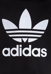 Bílé logo trefoilu Adidas se třemi pruhy nad názvem značky v malých písmenech na černém textilním pozadí.