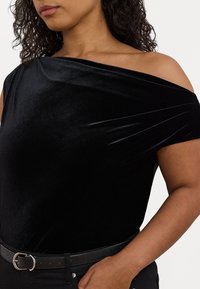 Svart sammet off-shoulder topp med draperad tygdetalj. Texturen är mjuk med en figurnära silhuett. Parat med ett svart bälte.