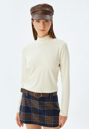 Crème geribbelde turtleneck, gecombineerd met een navy geruit minirok met gele accenten, en een bruine leren pet met een ronde rand.
