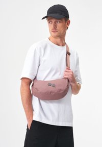 Helle rosa Schultertasche mit zwei grauen Knöpfen, aus weichem Stoff hergestellt. Abgerundete Form mit einem verstellbaren Tragegurt, der quer über den Körper getragen wird.