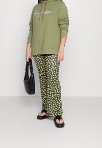 Olivgrüner Hoodie mit weißem Logo, schwarze und gelbe Blumenmusterhose, schwarze Tasche mit strukturiertem Finish und klobige schwarze Sandalen.