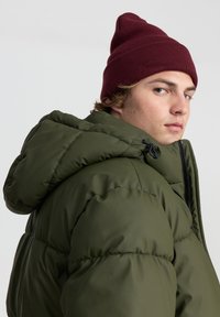 Giovane uomo che indossa un berretto di maglia color bordeaux e una giacca imbottita verde oliva con cappuccio, che guarda oltre la spalla contro uno sfondo neutro.