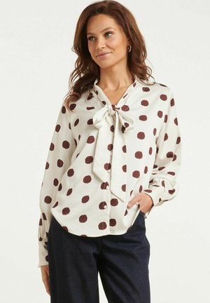 Vrouw met een witte blouse met bruine polkadots en een gestrikte strik bij de hals, gecombineerd met een donkere broek, staand met één hand in de zak.