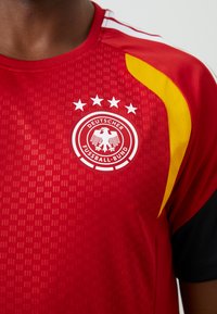 Κόκκινη αθλητική φανέλα με στρογγυλό γιακά, υφή υφάσματος, πλευρικές λεπτομέρειες σε κίτρινο και κεντρικό λογότυπο με το έμβλημα της Γερμανικής Ποδοσφαιρικής Ομοσπονδίας.
