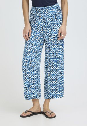 Kobieta ubrana w szerokie spodnie w niebiesko-biały wzór, czarny bezrękawnik i czarne klapki, stojąca na jednolitym tle.