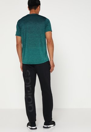 Mand iført en grøn gradient kortærmet skjorte og sorte Adidas sweatpants med logo på benet, stående mod en ensfarvet hvid baggrund.