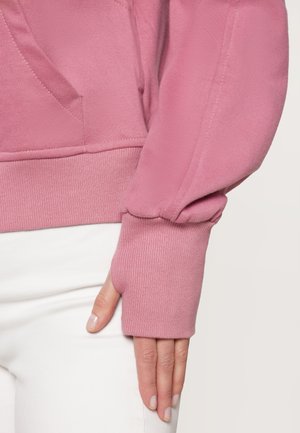 Rosa Sweatshirt mit geripptem Saum und Bündchen, ausgestattet mit einer Fronttasche und Daumenlöchern. Das Material scheint weich zu sein und wurde für Komfort entworfen.