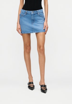 Modelka nosí světle modrou džínovou mini sukni s předními kapsami, černý tílkový top a černé nazouvací boty s kovovými přezkami a špičatou špičkou.