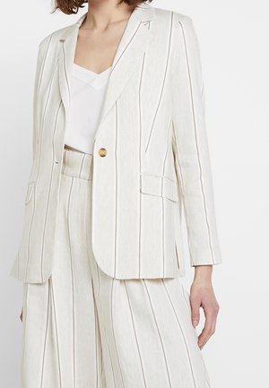 Vrouw die een crèmekleurig pak draagt met dunne verticale strepen, bestaande uit een blazer met één knoop en bijpassende wijde broek met hoge taille.