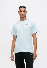 Gaiši zila kokvilnas T-kreklu ar apaļu kaklu, īsām piedurknēm un sarkanu Nike logo uz krūtīm. Valkāts ar melnām biksēm.