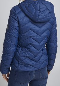 Giacca a vento blu con cappuccio, caratterizzata da un motivo trapuntato a chevron e polsini elasticizzati. Il materiale appare lucido e imbottito per il calore.