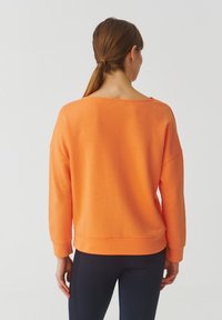 Felpa arancione con un taglio relaxed, scollo ampio, spalle cadenti e polsini a costine. Texture liscia, abbinata a leggings scuri.