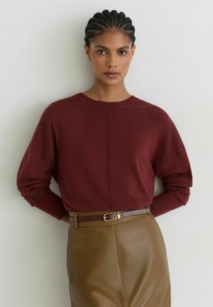 Kvinde med flettet hår iført en bordeaux sweater og olivengrøn lædernederdel med en brun bælte, stående mod en hvid væg.