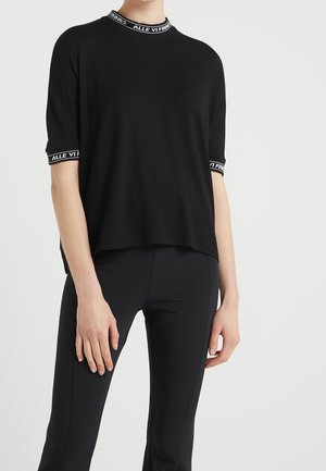 Persoon draagt een zwart kortärmelig shirt met witte tekst op de kraag en mouwboorden, gecombineerd met nauwsluitende zwarte broek tegen een witte achtergrond.