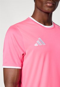 Ροζ αθλητική μπλούζα με στρογγυλή λευκή λαιμόκοψη, με υφασμάτινη υφή και λευκό λογότυπο Adidas στο αριστερό σ τήθος. Κοντά μανίκια.