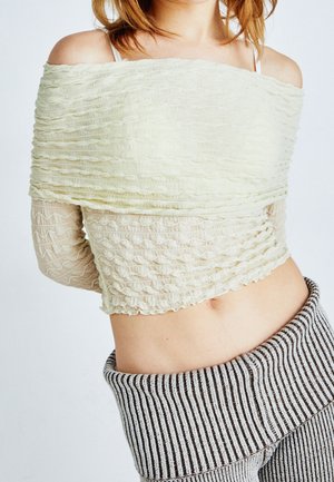 Ung kvinde iført en off-shoulder tekstureret cremefarvet top med lange gennemsigtige ærmer og højtaljede, ribstrikkede bukser i grå og sort stribet mønster.