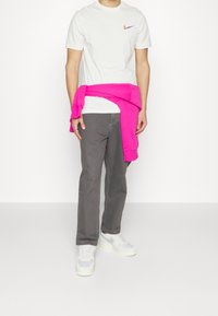 Vitt Nike-t-shirt med en färgglad logotyp, grå byxor och en rosa sweatshirt knuten runt midjan. Ljusgrå sneakers fulländar looken.