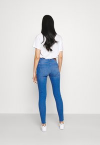 Vaqueros azules de tiro alto con un ajuste estrecho. Vista trasera con dos bolsillos, textura de mezclilla suave y un sutil desgaste en las piernas. Combinados con zapatillas blancas.