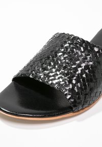 Sandale slide noire avec une bride en cuir tressé brillant et un lit de pied lisse, avec un bord de semelle marron clair.