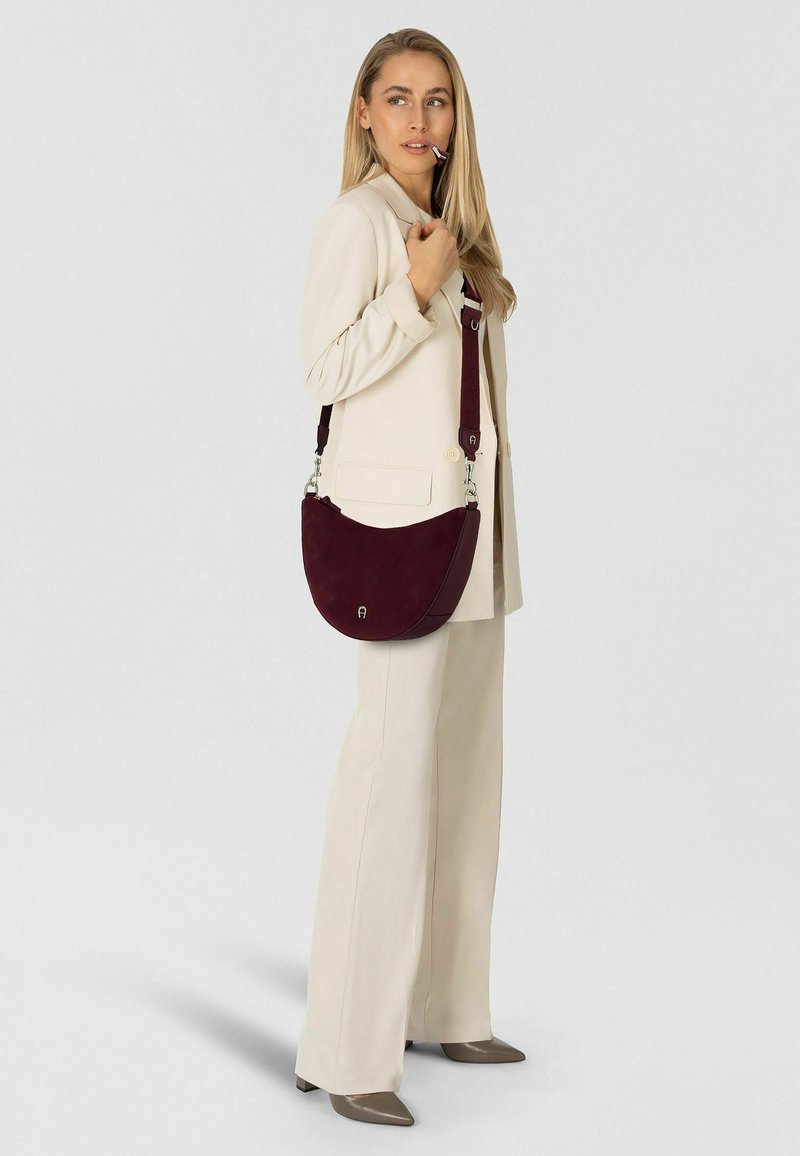 Donna in completo crema che tiene la tracolla di una borsa a tracolla bordeaux, guarda di lato con una penna in bocca su sfondo semplice.