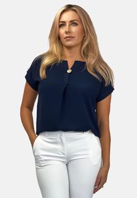 Granatowa bluzka z krótkim rękawem, dekoltem w serek i ozdobnym guzikiem. Połączona z białymi spodniami o dopasowanym kroju. Gładki tekstura materiału.