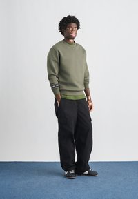 Σκοτεινό πράσινο φούτερ με ριμπ λαιμόκοψη, φορεμένο πάνω από ένα πιο ανοιχτό πράσινο πουκάμισο. Μαύροι παντελόνια cargo και μαύρα αθλητικά παπούτσια ολοκληρώνουν το ντύσιμο.