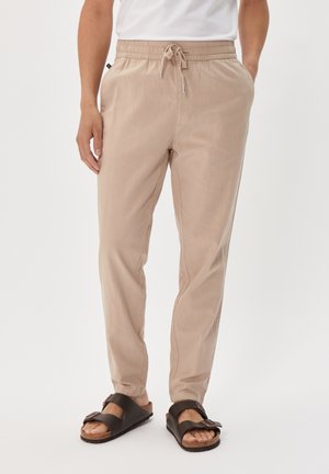 Mann trägt beige Kordelzughose mit Taschen und dunkelbraunen Schnallensandalen, Hände in den Taschen, steht vor weißem Hintergrund.