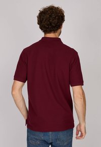 Polo shirt bordeaux avec col, manches courtes et fentes latérales. Fabriqué en tissu respirant, avec une texture lisse et un design minimaliste.