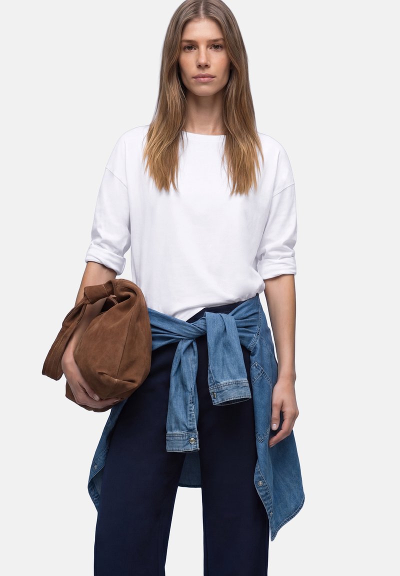 Femme aux cheveux longs portant un pull blanc et un pantalon foncé, une veste en jean nouée autour de la taille, tenant un grand sac marron dans la main gauche.