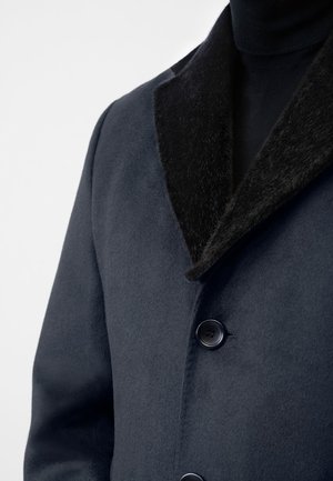 Marineblauwe wollen blend jas met een zwart nepbont kraag, voorzien van twee knopen en een zachte textuur, getoond van de kraag tot het borstgebied.