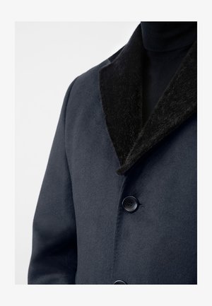 Mørkeblå uldblandet frakke med en sort faux fur-krave, som har to knapper og en blød tekstur, vist fra kraven og ned til brystområdet.
