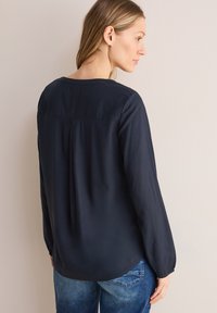 Marineblauwe blouse met lange mouwen, rond decolleté, subtiele plooival aan de achterkant en een soepele textuur, gecombineerd met lichtblauwe denim jeans.