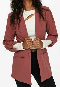 Blazer rose poudré à manches longues, avec deux poches avant et une coupe ajustée. En dessous, un haut blanc avec un design ajouré.