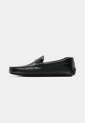 Black leather loafers with a glossy finish, rounded toe, and decorative hardware on the front. Low-profile rubber sole. Smooth texture.
