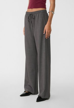 Grijze wijde broek met een ontspannen pasvorm, voorzien van een tailleband met trekkoord, een soepele textuur en een subtiel ruitpatroon. Zwarte puntige schoenen accentueren de look.