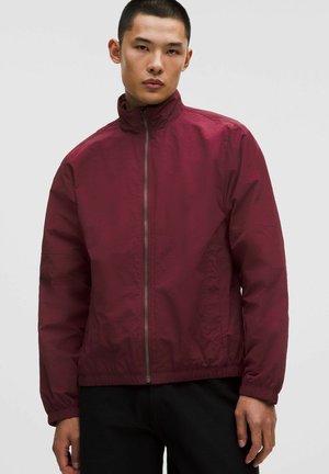 Giovane uomo che indossa una giacca bordeaux con zip e collo alto, pantaloni neri, in piedi davanti a uno sfondo chiaro e uniforme.