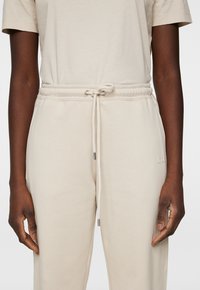 Beige Jogginghosen mit elastischem Bund, Kordelzugverschluss und dezentem Logo-Detail an der linken Hüfte. Weiche, glatte Baumwollqualität.