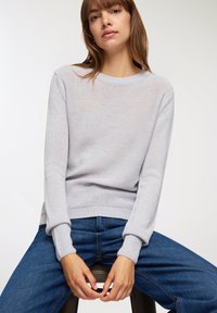 Donna con capelli castani lisci che indossa un maglione lavorato a maglia grigio chiaro e jeans blu, seduta su uno sgabello con le mani intrecciate.