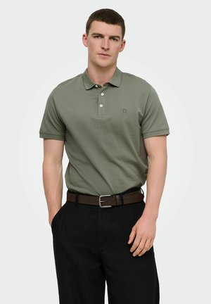 Mladić u maslinasto zelenoj polo majici kratkih rukava, utrgnutoj u crne hlače s smeđim remenom, stoji s jednom rukom u džepu.