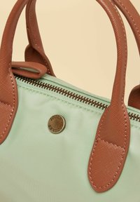 Bolso de mano verde claro con asas marrones texturizadas, un botón de latón grabado con "Joules" y una cremallera marrón sobre un fondo beige.