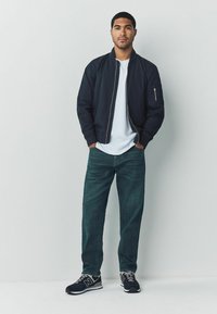 Mereväe pommituslendurijope, valge T-särk, tikitud sinaka tooniga kulunud teksad, mustad tossud hallide detailidega, modell seisab käed taskutes.