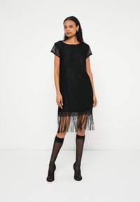 Melnas mežģīņu kleita ar īsām piedurknēm, bārkstīm apakšmalā un brīva piegriezuma. Valkāta kopā ar melnām augstām zeķēm virs ceļa un atklāta purngala augstiem papēžiem.