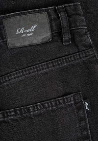 Bolsillo trasero de jeans de mezclilla negra y presilla del cinturón mostrando la etiqueta de la marca Reell cosida en la pretina y una pequeña etiqueta de tela negra en el borde del bolsillo.