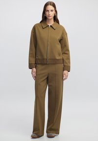 Chaqueta marrón con cremallera y un cuello amplio, acompañada de pantalones sueltos a juego. Tejido suave con puños y cinturilla de canalé. Diseño sencillo.