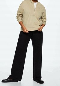 Personne portant un pull beige à demi-fermeture éclair, un pantalon noir à jambes larges et des chaussures noires sans lacets, debout devant un fond clair uni.