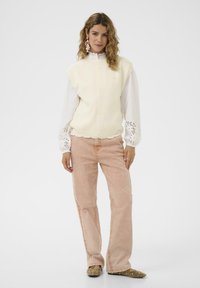 Vaaleanbeige neuleliivi, jossa on kaareva helma, valkoisen paidan päällä, yhdistettynä vaaleanpunaisiin leveälahkeisiin housuihin ja beigeihin koristelluihin kenkiin.