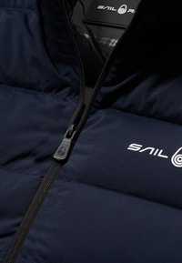 Marinefarbender gepolsterter Weste mit hohem Kragen, ausgestattet mit einem schwarzen Reißverschluss und dem weißen "Sail"-Logo. Glattes, glänzendes Material mit gesteppten Paneelen.