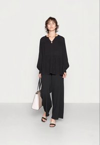 Top preto de mangas longas com decote em V e design plissado, combinado com calças largas pretas. Saco tote em tom claro e sandálias pretas.