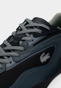 Basket Lacoste avec une tige synthétique noire et vert foncé mélangée, des lacets gris, un matériau texturé et un logo bien visible sur le côté.