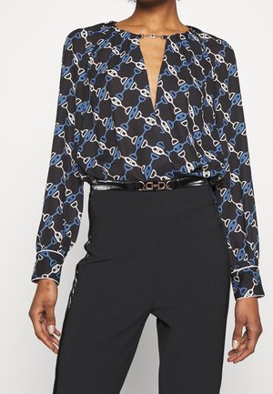 Kobieta w czarnej bluzce z wzorem z białych i niebieskich połączonych łańcuchów, w zestawieniu z wysokim stanem czarnych spodni i błyszczącym czarnym paskiem.